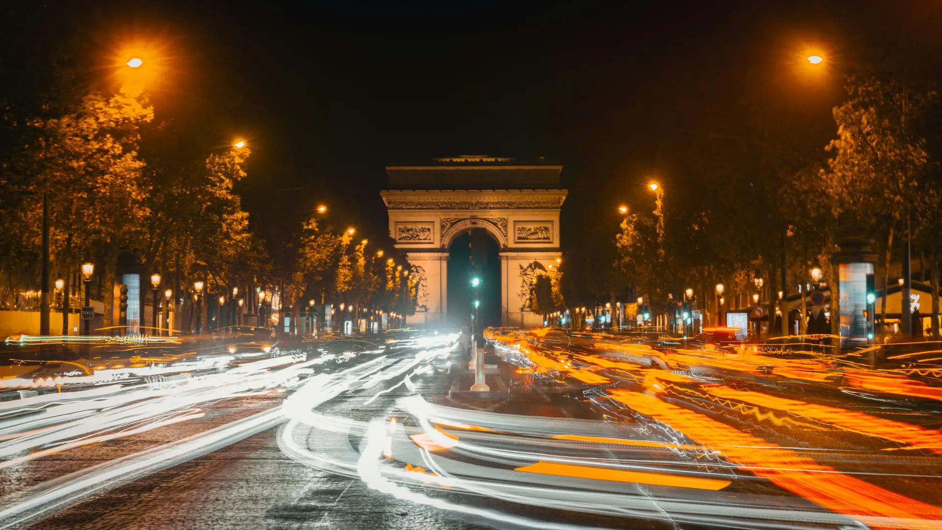 Car light trails streaming along a tree-lined avenue at night toward the floodlit Arc de Triomphe