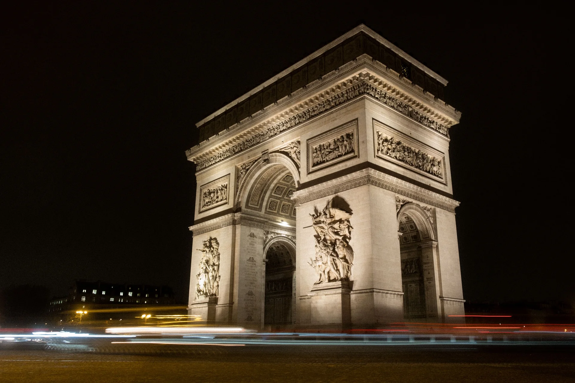 The Arc de Triomphe floodlit at night, its carved pillars bright against a black sky, with car light trails streaking across Place Charles de Gaulle